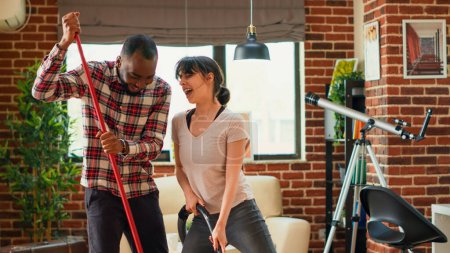 Téléchargez les photos : Interracial couple showing dance moves and having fun spring cleaning, mopping and vacuuming wooden floors in living room. Happy life partners enjoying using mop and vacuum cleaner. - en image libre de droit