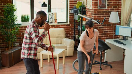 Photo for Smiling young people using tools to clean household, listening to music and dancing while they use mop and vacuum. Diverse couple cleaning apartment floors, sweeping dust with solution. - Royalty Free Image