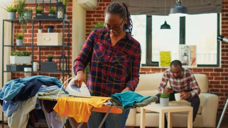 Téléchargez les photos : African american girl using steam on ironing board to do domestic work, casual girlfriend ironing laundered clothes in living room. Young woman smoothing out garment and clothing, chores. - en image libre de droit