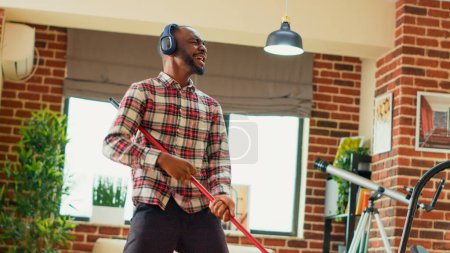 Téléchargez les photos : African american man dancing and using mop to wash floors, sweeping dust or dirt in living room. Young male adult having fun cleaning apartment, mopping with washing solution. Handheld shot. - en image libre de droit