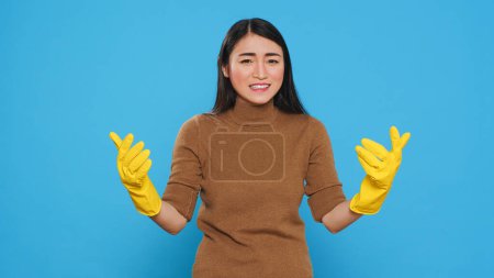 Téléchargez les photos : Nervous asian housekepper with protective gloves having problem with house cleaning, standing with arm crossed in studio over blue background. Maid having negative expression - en image libre de droit