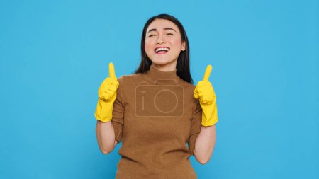Téléchargez les photos : Positive joyful maid smiling while doing ok gesture after finishing to clean client house, posing in studio over blue background. Cheerful housekeeper is responsible for providing cleaning services - en image libre de droit