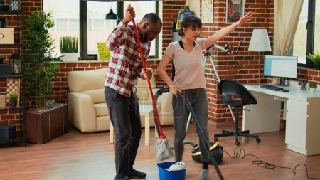 Foto de Cheerful people showing dance moves and having fun cleaning living room, using mop to sweep dust and vacuum at home. Couple dancing and singing, using floor cleaner, spring cleaning. - Imagen libre de derechos