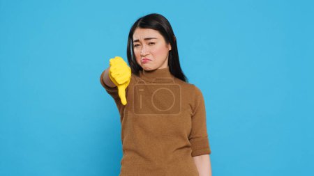 Photo for Upset asian maid doing thumbs down gesture while cleaning client house using houseclean detergent, posing in studio. Housekeeper is also knowledgeable about proper hygiene and cleanliness practices - Royalty Free Image
