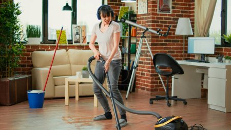 Téléchargez les photos : Female adult listening to music on headset and vacuuming floors at home, doing spring cleaning work in apartment. Young wife singing and doing dance moves while she uses vacuum. - en image libre de droit
