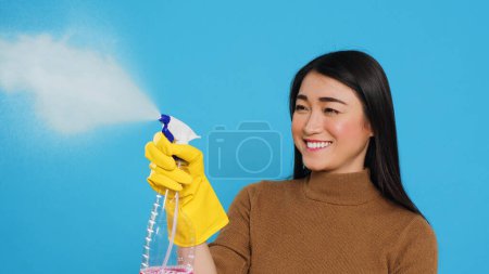 Téléchargez les photos : Housekeeper wearing yellow rubber gloves while holding spray and duster during houseclean, standing over blue background. Housewife primary responsibilities is to maintain cleanliness and hygiene - en image libre de droit