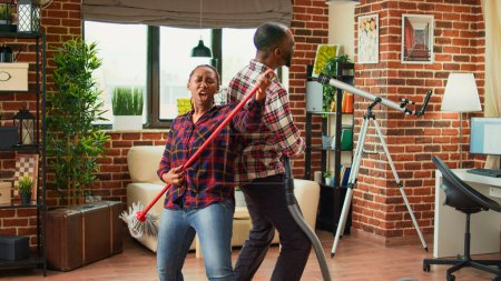 Photo for Smiling couple dancing and cleaning apartment with music, showing dance moves and having fun. Life partners feeling happy while they clean wooden floors with mop and vacuum cleaner. - Royalty Free Image