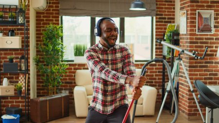 Photo for Cheerful funny boyfriend using mop to clean wooden floors, cleaning household and listening to music on headphones. Young smiling man enjoying mopping to clean dirt, household chores. - Royalty Free Image