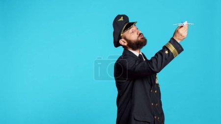 Téléchargez les photos : Homme caucasien habillé en pilote jouant avec un jouet d'avion, montrant un petit avion artificiel miniature à la caméra. Jeune adulte travaillant comme commandant de bord tenant un faux mini-avion sur fond bleu. - en image libre de droit
