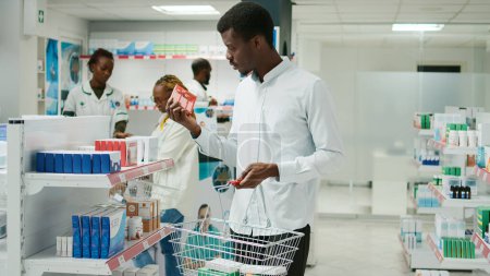 Téléchargez les photos : African american customer putting medicaments in basket, looking for vitamins and bottles of pills. Young male client buying prescription treatment and supplements, pharmacy shop. - en image libre de droit