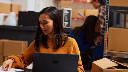 Photo for Asian employee working on stock logistics with laptop, planning order shipment for business development in warehouse. Woman doing supply chain management at desk, retail goods. Handheld shot. - Royalty Free Image