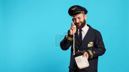 Photo for Commercial pilot talking on landline phone call in studio, answering office telephone with cord for remote conversation. Young smiling airliner in uniform chatting on phone line. - Royalty Free Image