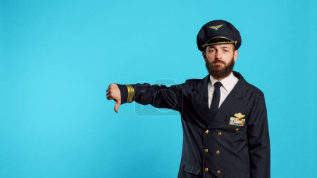 Téléchargez les photos : Professional aviator showing thumbs down sign, expressing dislike and bad negative gesture. Airline pilot in uniform doing disapproval and failure symbol, commercial flights occupation. - en image libre de droit