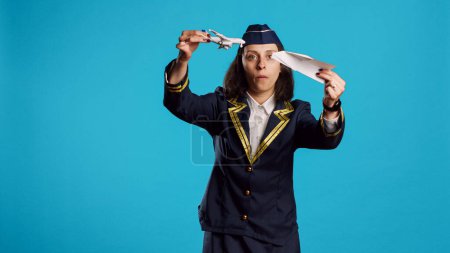 Photo for Positive stewardess playing with toy and paper plane, having fun with origami and mini artificial airplane in studio. Happy air hostess wearing flying uniform working in aviation industry. - Royalty Free Image