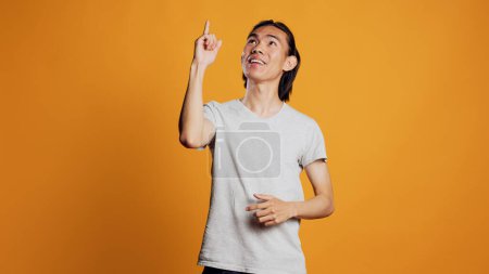 Téléchargez les photos : Young adult pointing above head with index fingers, looking up with confidence in studio. Smiling guy indicating upwards direction over orange background, friendly casual person. - en image libre de droit