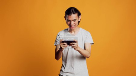 Photo for Happy male model playing video games on smartphone, using online gaming competition to have fun in studio. Young asian guy enjoying mobile game on phone, shooter challenge play. - Royalty Free Image