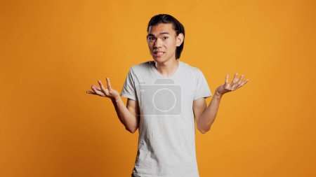 Foto de Clueless young person doing i dont know sign in studio, acting uncertain and unsure about answer to question. Asian guy being carefree and casual, acting doubtful over orange backdrop. - Imagen libre de derechos