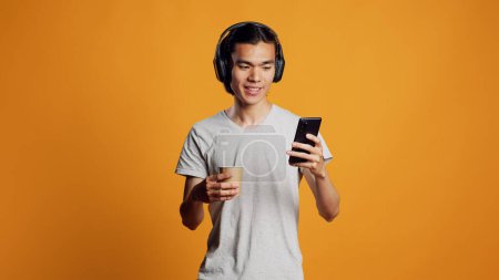 Téléchargez les photos : Modern person listening to music and using smartphone, having fun with mp3 songs on headphones. Young asian man enjoying sound with audio headset, checking smartphone app on camera. - en image libre de droit