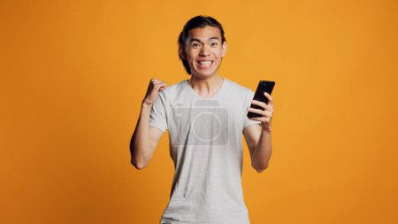 Téléchargez les photos : Heureux asiatique gars navigation sur Internet sur téléphone mobile, en utilisant un smartphone pour navigateur de médias sociaux sur la caméra. Personne souriante insouciante vérifiant le site Web en ligne sur fond orange. - en image libre de droit