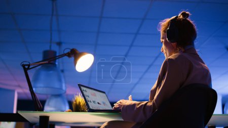 Photo for Executive assistant listening to music late at night, wearing headset and enjoying work. Young employee with heeadphones analyzing financial graphs report with analytics. Handheld shot. - Royalty Free Image