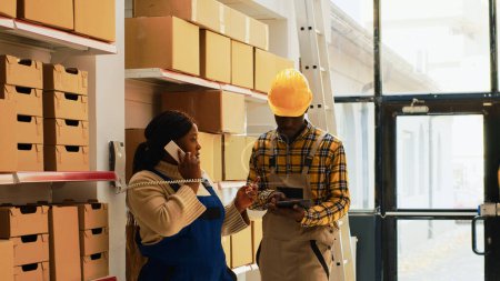 Foto de Depot manager answering phone call in warehouse space, preparing new supplies order with male employee. Young man writing list of goods on clipboard files, supervisor using landline phone. - Imagen libre de derechos