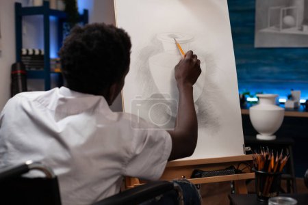 Photo for Drawing professional artist who uses wheelchair to get around starting pencil outline of vase in home art studio. Young african american man with artistic abilities spending time sketching at house. - Royalty Free Image
