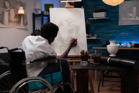 Téléchargez les photos : Young african american man with artistic abilities living with health condition making pencil sketch of vase at home art studio. Male wheelchair user spending time doing drawing task. - en image libre de droit