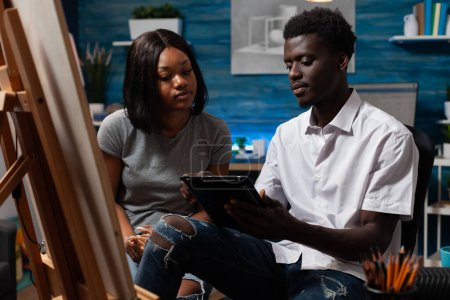 Téléchargez les photos : African american friends with creative abilities taking online distance art classes from home with electronic tablet. Artists looking for references to create drawing artwork on canvas easel. - en image libre de droit