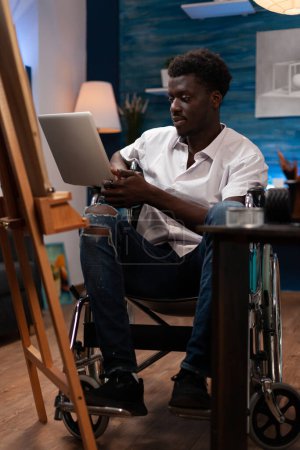 Foto de Person with artistic skills and disabilities taking online drawing courses with laptop. African american wheelchair user looking for inspiration on art web sites to create sketch masterpiece at - Imagen libre de derechos
