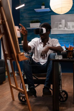Téléchargez les photos : African american male artist wheelchair user visualizing digital art references within metaverse to draw. Young art student looking for inspiration inside virtual universe with vr glasses. - en image libre de droit