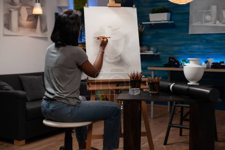 Foto de Profesora profesional afroamericana de artes del dibujo que crea una obra maestra de dibujo a lápiz de jarrón sobre caballete de tela, sentada en casa dibujando un taller con herramientas creativas al lado. - Imagen libre de derechos