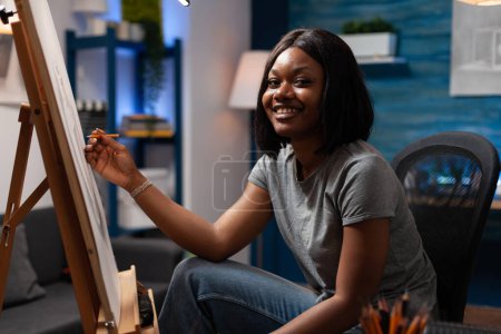 Téléchargez les photos : African american woman with artistic skills happily drawing on blank canvas easel in house made art studio. Female sketch student learning to draw by hand with pencils in atelier at home. - en image libre de droit