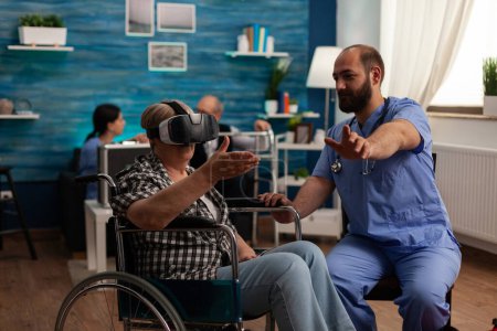 Foto de Elderly woman in wheelchair using virtual reality glasses in common room of nursing home, male nurse providing assistance and teaching how to use modern technology. Time for fun in care center. - Imagen libre de derechos