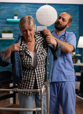 Téléchargez les photos : Infirmière aidant une patiente en maison de retraite. Femme âgée utilisant marchette pour le soutien à la marche tandis que le spécialiste l'aide à mettre sur la veste après la séance de physiothérapie. - en image libre de droit