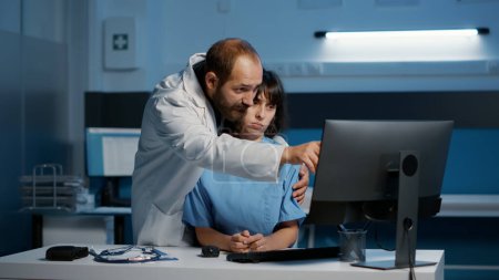 Photo for Nurse standing at desk typing medical expertise on computer while physician doctor touching her inappropriately during over night shift in hospital office. Assistant suffering from emotional trauma - Royalty Free Image
