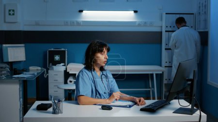 Téléchargez les photos : Physician nurse with blue uniform and stethoscope analyzing patient disease report working at health care treatment late at night in hospital office. Assistant checking medical expertise on computer - en image libre de droit