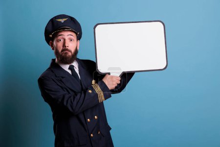 Photo for Surprised airplane pilot holding white empty speech bubble with copy space, aviator with communication frame mock up. Plane captain in uniform standing with blank message balloon - Royalty Free Image