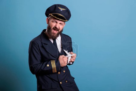 Photo for Funny pilot in uniform playing with small airplane model, aviation academy aviator holding commercial passenger plane toy. Aircraft crew member looking at camera, studio medium shot - Royalty Free Image