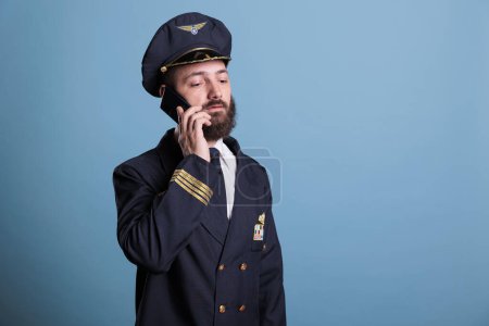 Photo for Middle aged pilot talking on smartphone, holding mobile phone, having conversation. Airlane captain in uniform with calm neutral facial expression answering telephone call - Royalty Free Image