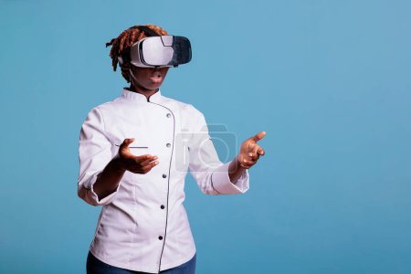 Photo for Serious professional chef using VR headset to explore augmented reality on blue studio background. Modern cook in uniform watching at the simulation of new cooking techniques. - Royalty Free Image