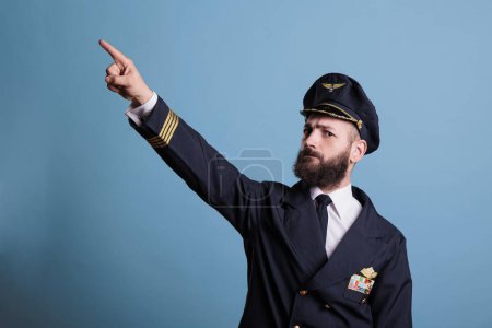 Photo for Serious captain pointing at sky with index finger, wearing professional aviation uniform, plane pilot looking at camera. Confident aviation academy aviator standing, studio medium shot - Royalty Free Image