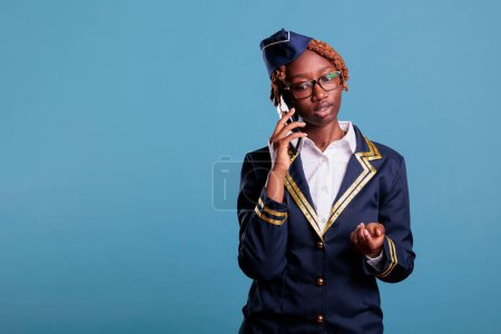 Photo for Concentrated female flight attendant talking on the phone dressed in uniform and eyeglasses. Stewardess looks serious while using mobile device in studio shot against blue background. - Royalty Free Image