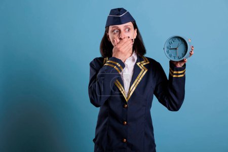 Photo for Anxious stewardess holding retro alarm clock, running late at airport, front view studio medium shot. Confused flight attendant overslept, time management, deadline, schedule concept - Royalty Free Image