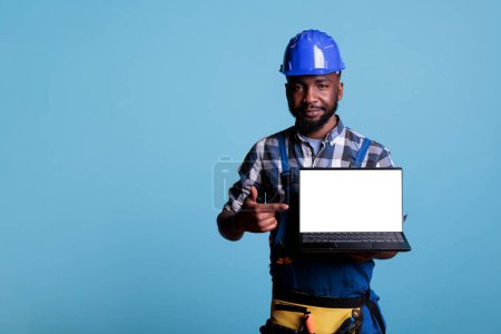 Photo for Smiling african american construction worker pointing at empty laptop screen with copy space, showing advertising mock up. Cheerful employee holding notebook, wearing coveralls in studio shot. - Royalty Free Image