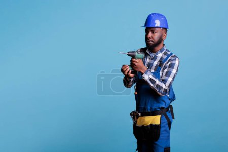 Foto de Concentrated african american builder holding cordless drill against blue background. Professional man carrying belt construction tools wearing hard hat and coverall, contrator job. - Imagen libre de derechos
