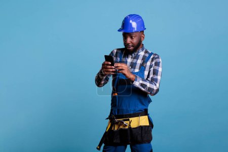 Téléchargez les photos : African american builder looking with amazed expression at cell phone screen, wearing coveralls and hard hat. Electrician with tool belt using mobile device against light blue background. - en image libre de droit