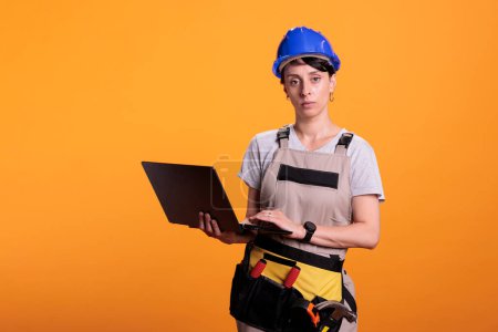 Photo for Female construction worker in overalls using laptop in studio to find renovating inspiration, looking at online website app on portable pc. Working with computer browsing internet. - Royalty Free Image