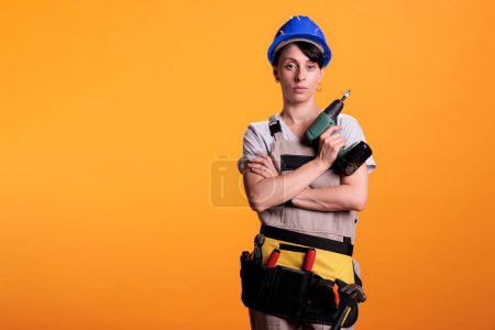 Photo for Engieering supervisor holding electric screw gun for construction industry advertisement in studio. Female builder worker in overalls and hard hat standing on yellow background with power drill. - Royalty Free Image