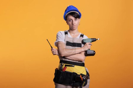 Photo for Angry construction worker holding drill gun and screwdriver, posing in studio. Woman dressed as building worker holding renovating tools and power screw drilling gun, serious builder. - Royalty Free Image