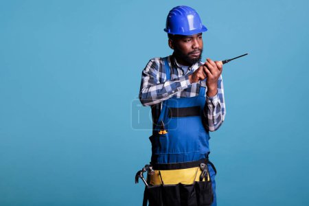 Téléchargez les photos : African american construction professional using screwdriver against blue background in studio portrait. Electrician doing electrical installation on wall dressed in uniform, hard hat and tool belt. - en image libre de droit